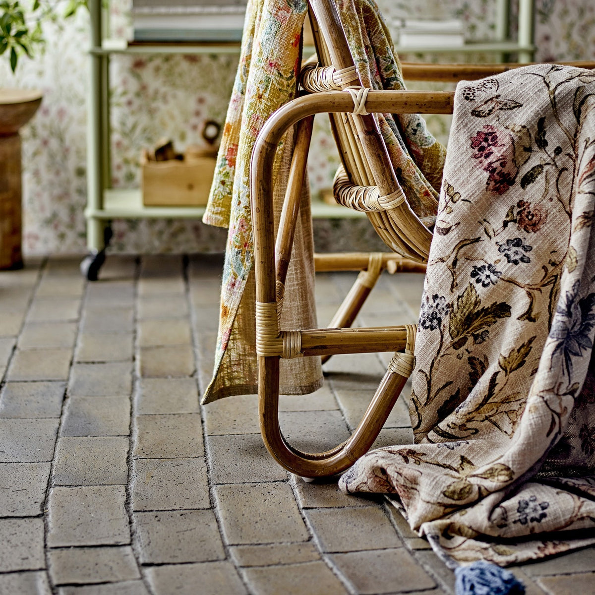 Yoti Floral Cotton Throw in multi draped over a rattan chair frame, showing its all-over floral print with trailing leaves and a tasselled corner against a tiled floor.
