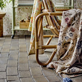 Yoti Floral Cotton Throw in multi draped over a rattan chair frame, showing its all-over floral print with trailing leaves and a tasselled corner against a tiled floor.