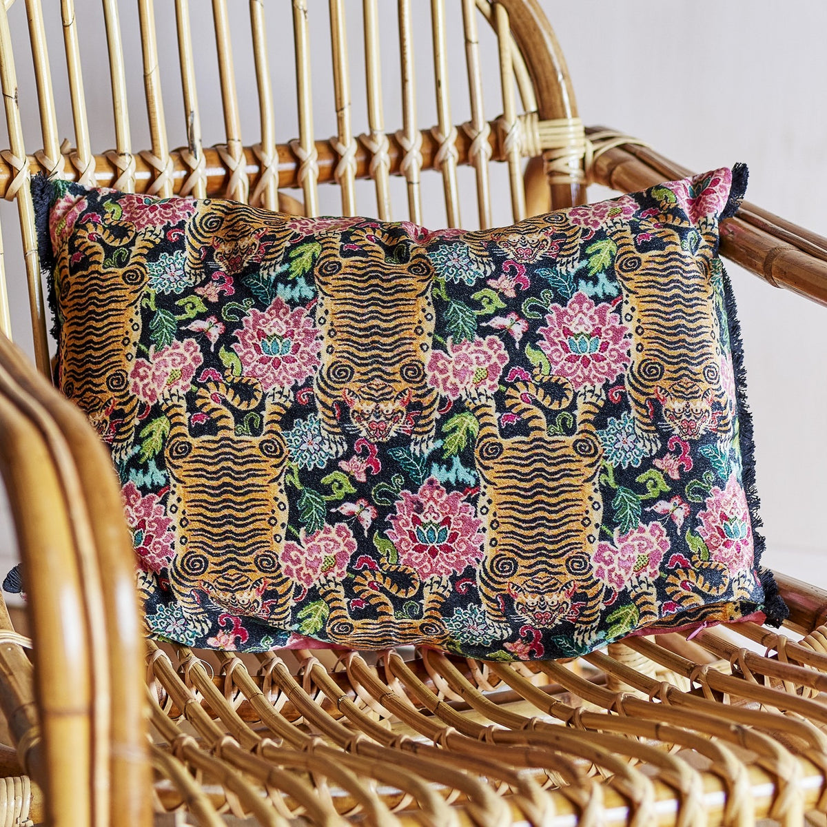 Lester Cotton Cushion in Multi, a rectangular lumbar cushion with a printed tiger and floral pattern in black, gold, and pink, shown resting on a woven rattan chair.