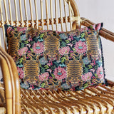 Lester Cotton Cushion in Multi, a rectangular lumbar cushion with a printed tiger and floral pattern in black, gold, and pink, shown resting on a woven rattan chair.