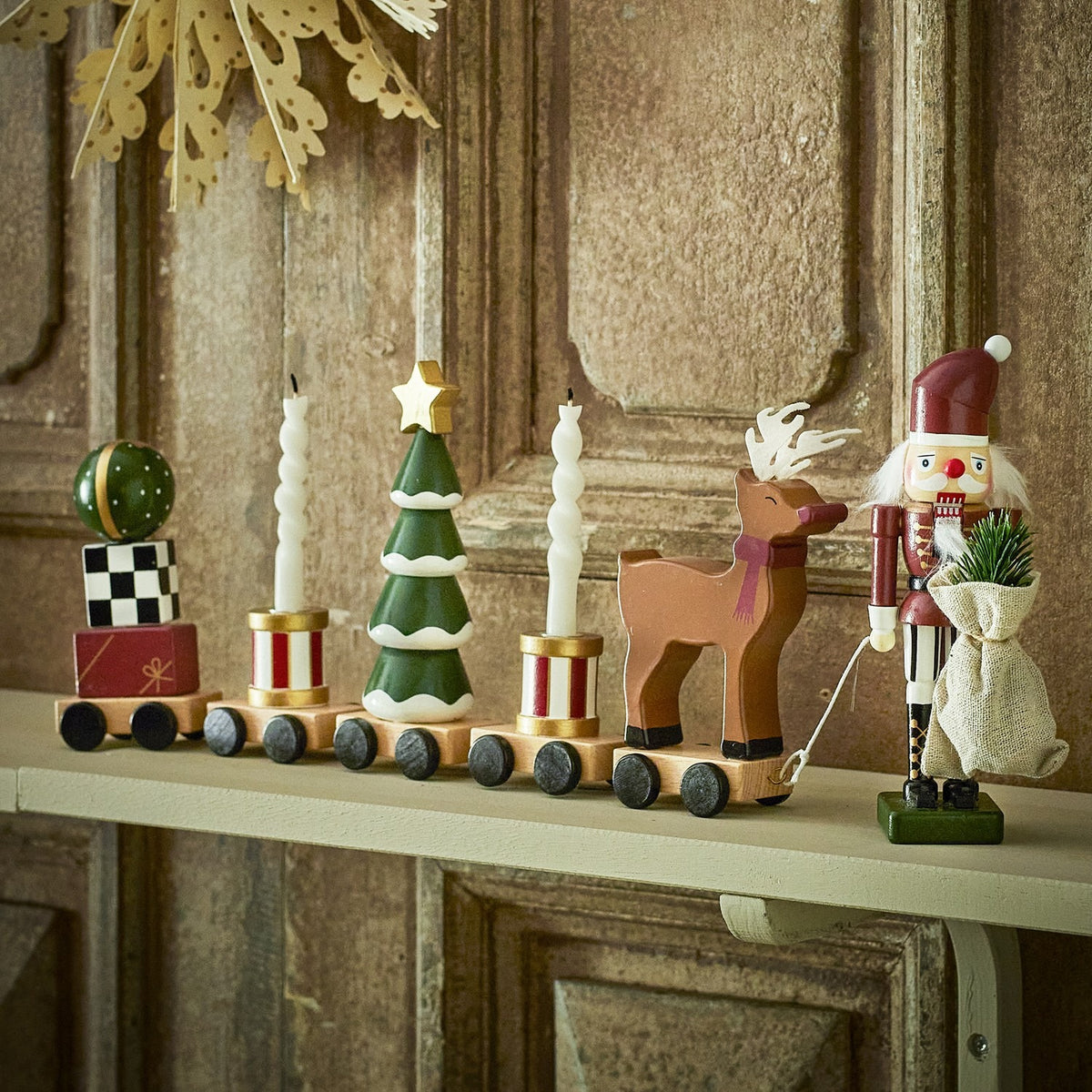 Vadim Christmas Decoration wooden train on a mantel shelf, with wagons carrying a bauble on presents, candle-topped drums, a snow-tipped tree, and a reindeer, led by a nutcracker figure.