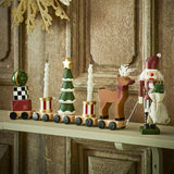 Vadim Christmas Decoration wooden train on a mantel shelf, with wagons carrying a bauble on presents, candle-topped drums, a snow-tipped tree, and a reindeer, led by a nutcracker figure.
