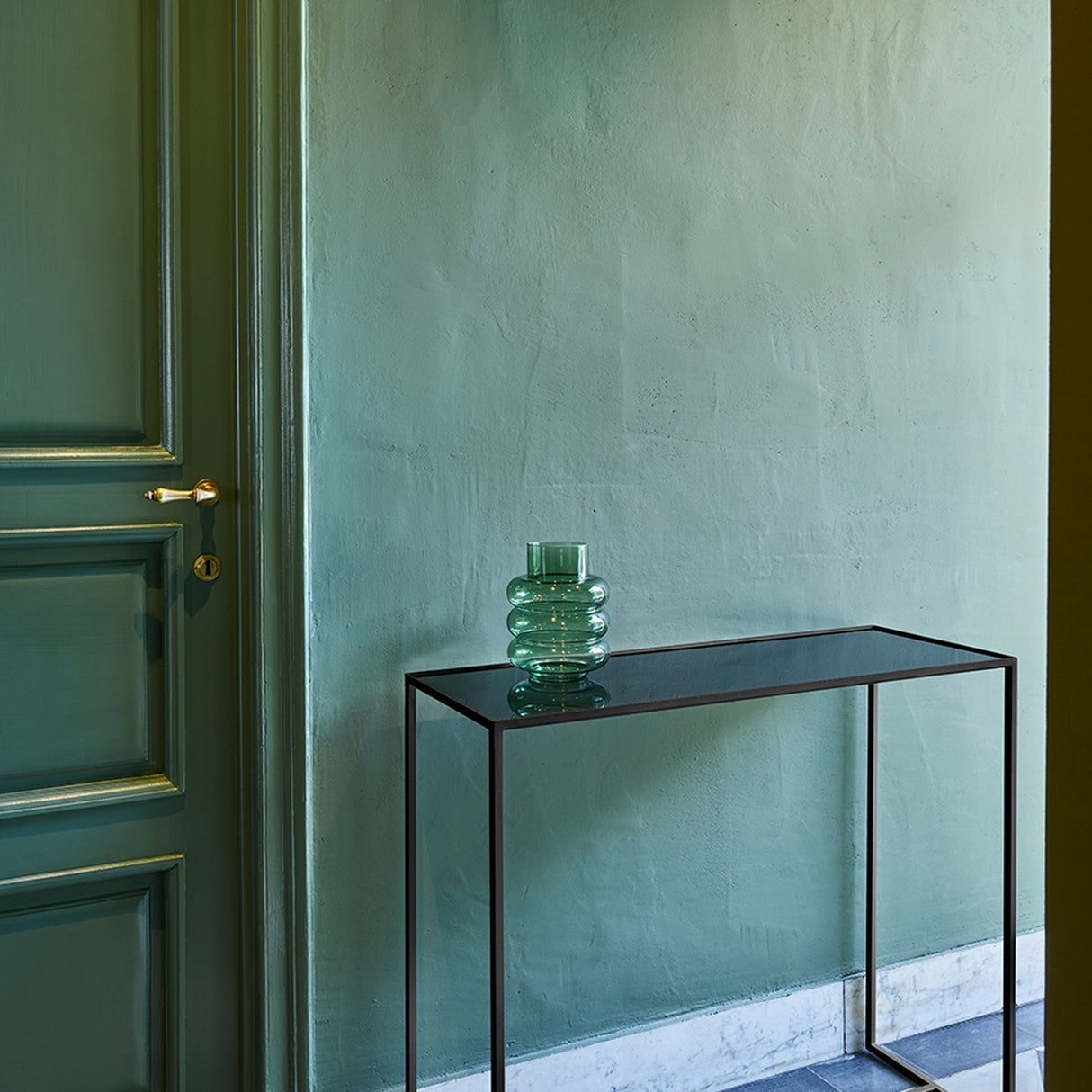 The mirrored console table against a deep green panelled wall in a hallway, with a green glass vessel standing on the top.