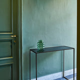 The mirrored console table against a deep green panelled wall in a hallway, with a green glass vessel standing on the top.