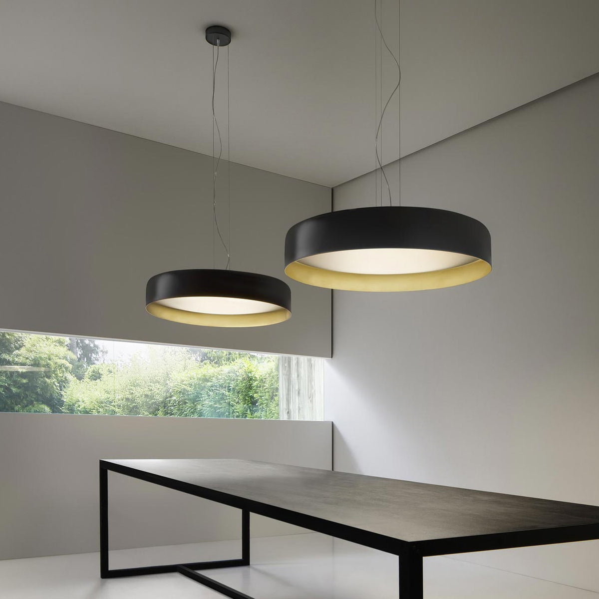 Ginevra 80cm LED pendant lights in matt black with brass inner rim and white diffuser, hung above a dark dining table in a modern grey interior.