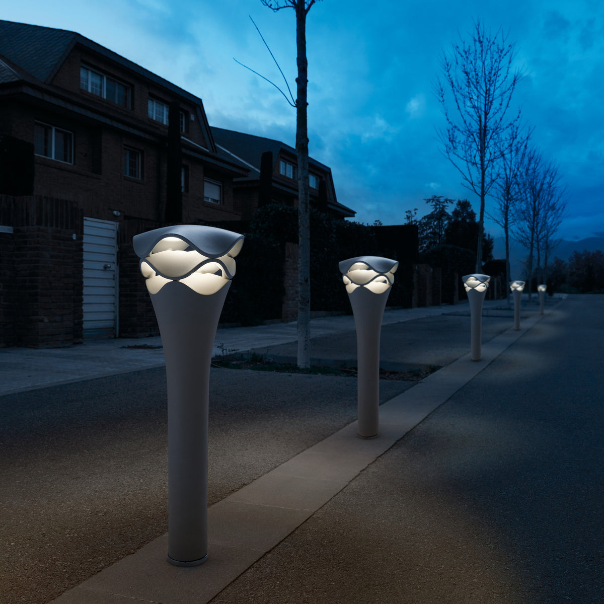 Cornet B/77 stone grey LED bollard lights lining a garden pathway at dusk, each with a slim flared stem and tiered wavy head glowing warm white light.