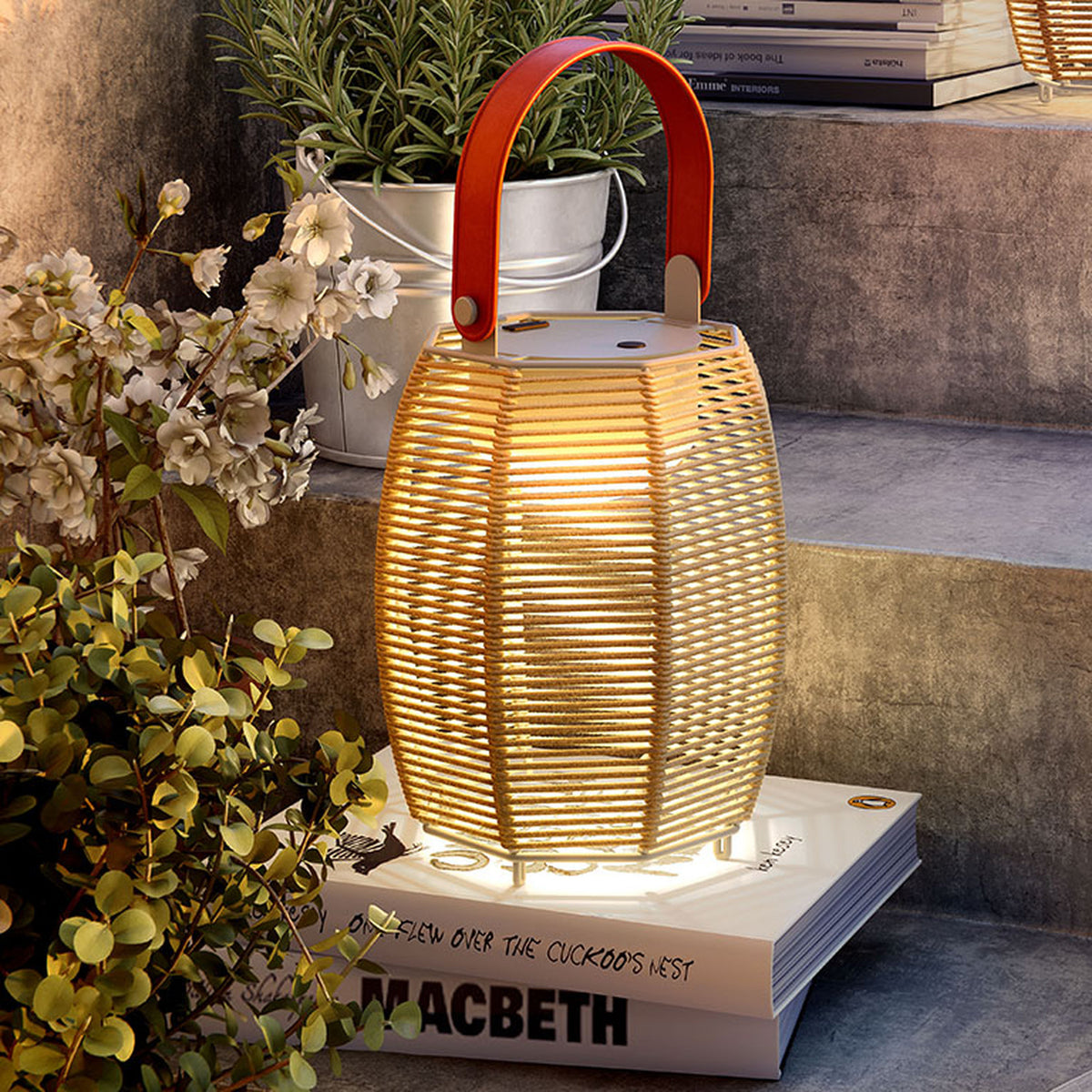 Tanit M/29/R rechargeable table lamp with woven brown fabric lantern shade, red carry handle, and integrated LED, shown outdoors on a book atop stone steps.