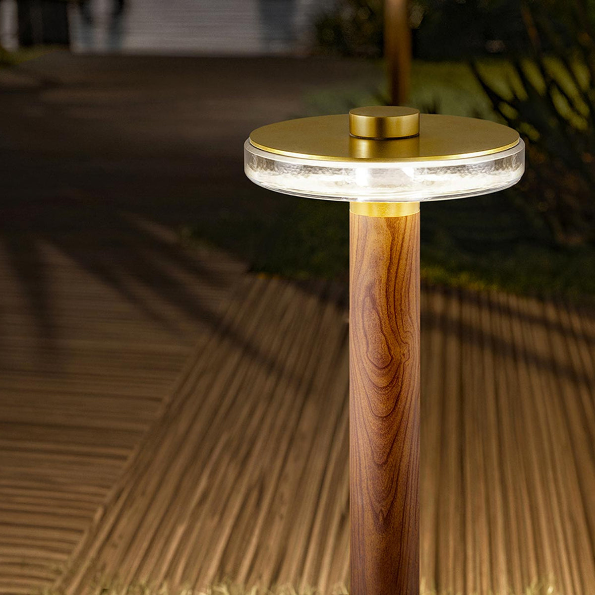 Venexia Glass LED Outdoor Floor Lamp with a wood column and round matt brass base, topped by a clear glass disc head glowing warm white, shown on a decked outdoor path at night.