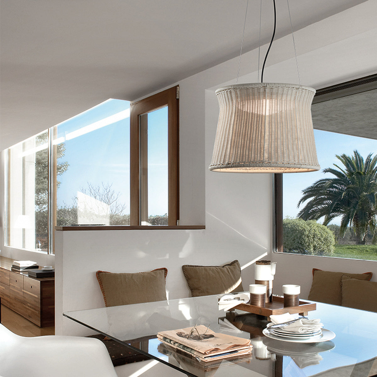 Syra 60 Aluminium LED Outdoor Pendant Light with a woven ivory white drum shade, narrow waist and flared rim, hung on fine wires above a glass dining table.