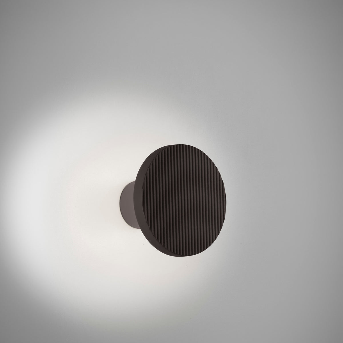 Arid A/01 round LED wall light in Textured Black aluminium, a fluted disc on a short stem casting a soft halo of light onto a plain grey wall.