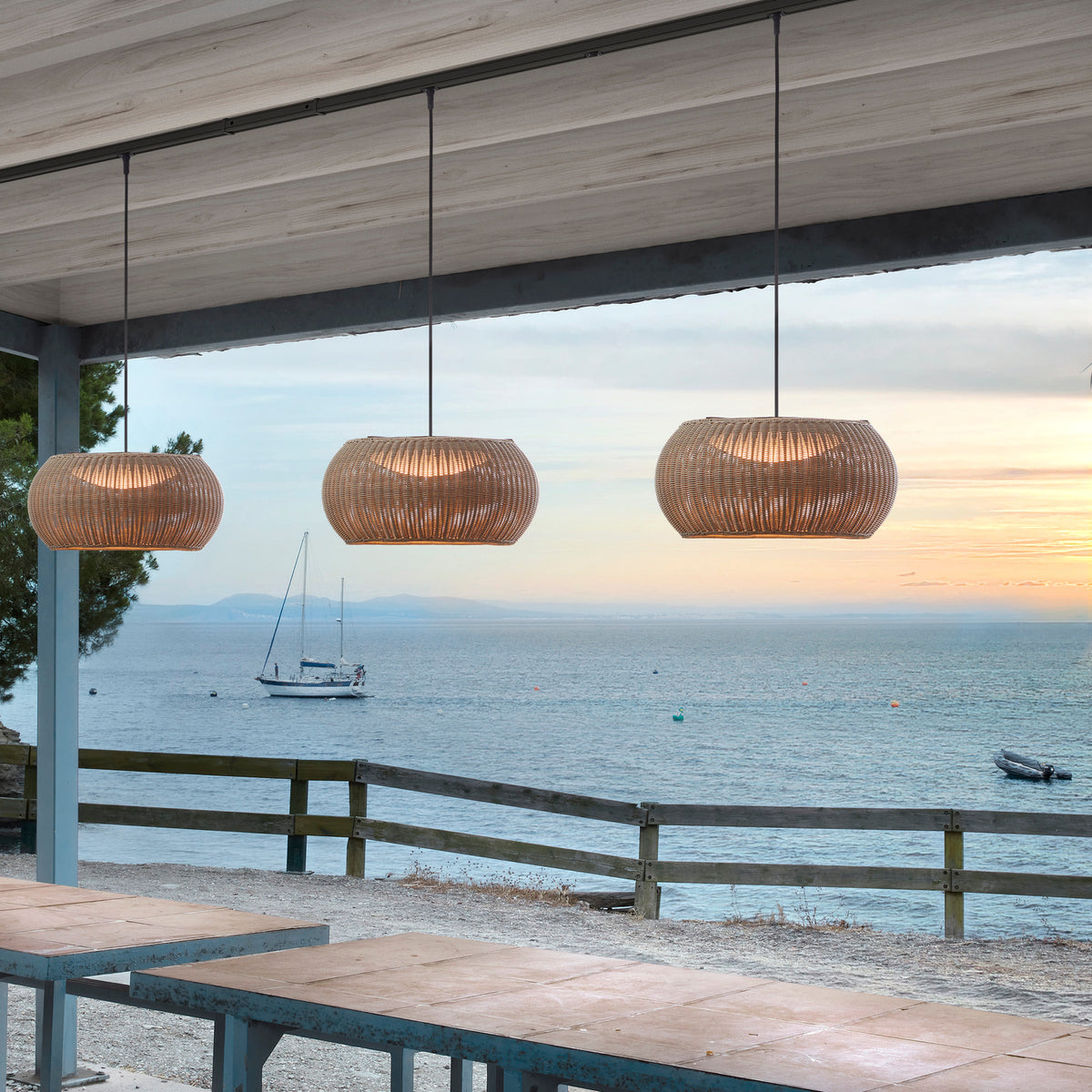 Three Perris S/47 Outrack pendant lights with woven brown globe shades hanging over a wooden outdoor table on a seaside terrace at dusk.