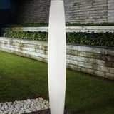 Maxi P/180 outdoor floor lamp, tall white polyethylene cylinder with fine vertical ribs, illuminated on a garden lawn beside wooden retaining walls and hedges at night.