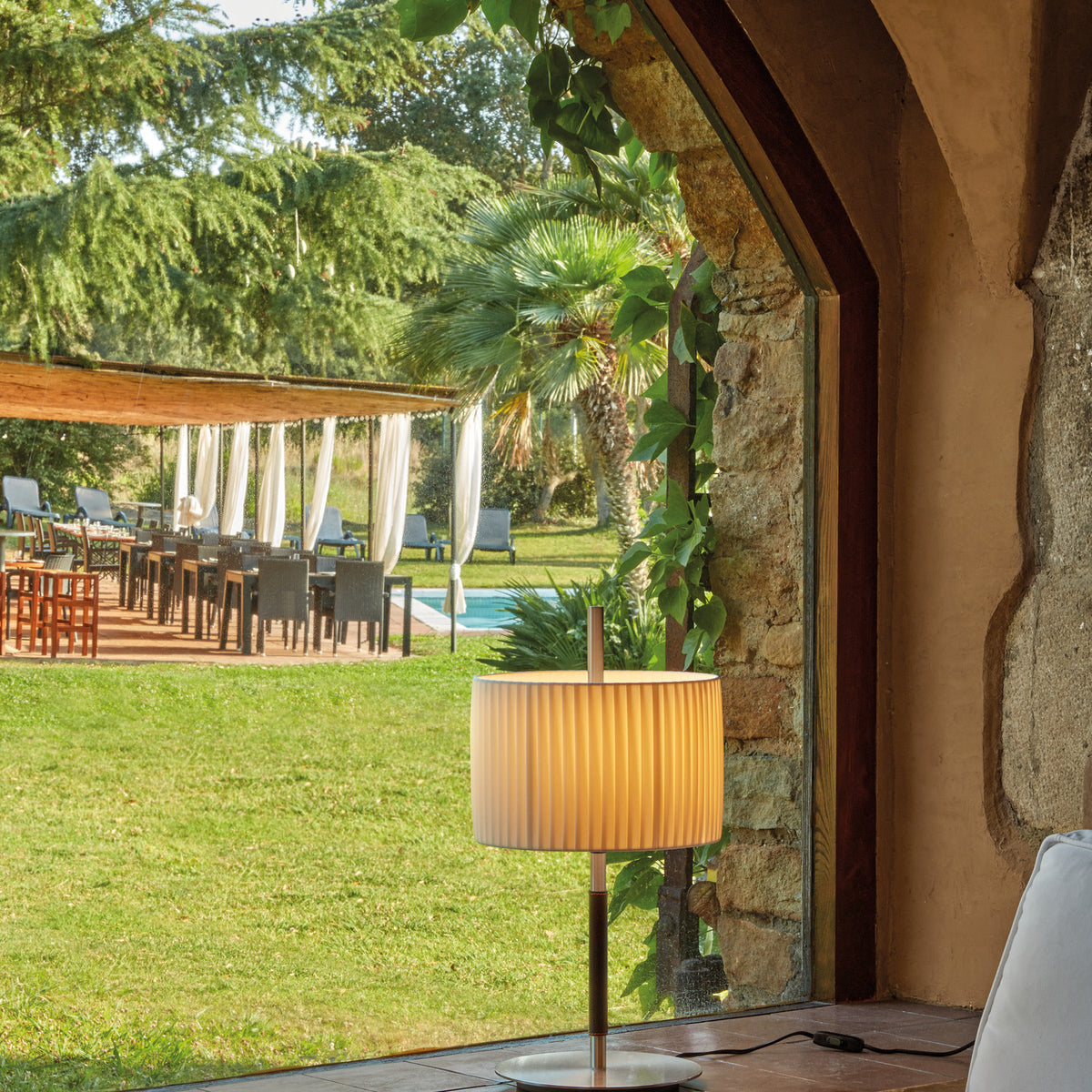 Danona T Ribbon Drum Table Lamp with pleated white fabric shade on a slim satin nickel stem and round base, shown on a terrace table by a glass archway.