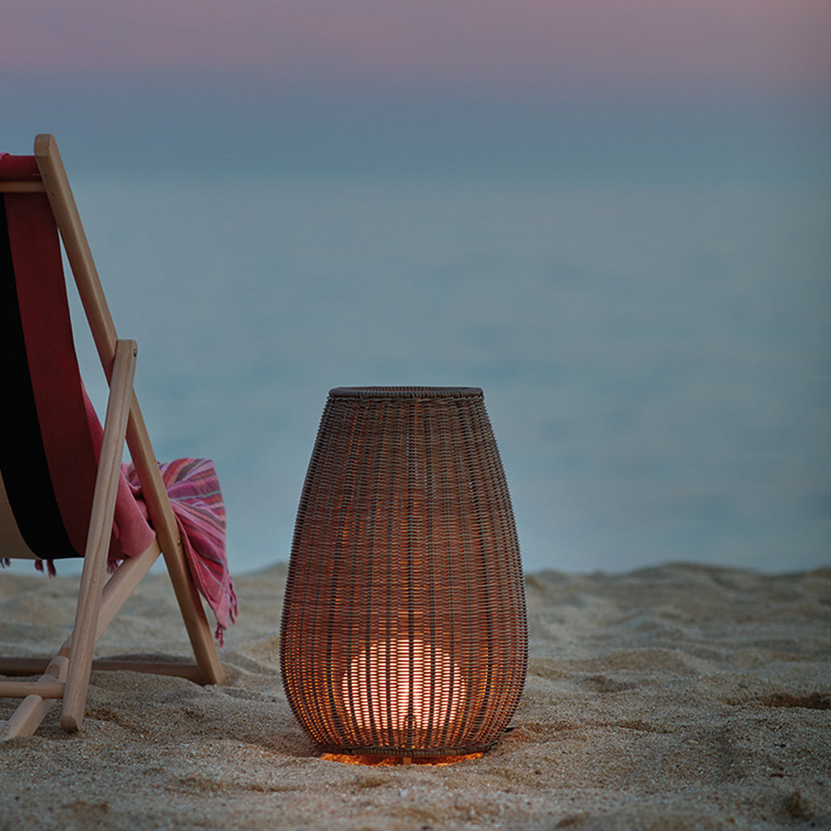 Amphora Woven Linear LED Outdoor Floor Lamp in beige, a tapering woven shade glowing warm white on sandy ground beside a deck chair at dusk.
