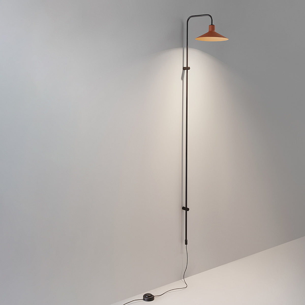 Platet A/05 Aluminium LED Wall Light with a tall slim black rod rising from floor level, curving at the top to hold a shallow terracotta cone shade, shown against a plain grey wall.