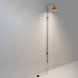 Platet A/05 Aluminium LED Wall Light with a tall slim black rod rising from floor level, curving at the top to hold a shallow terracotta cone shade, shown against a plain grey wall.