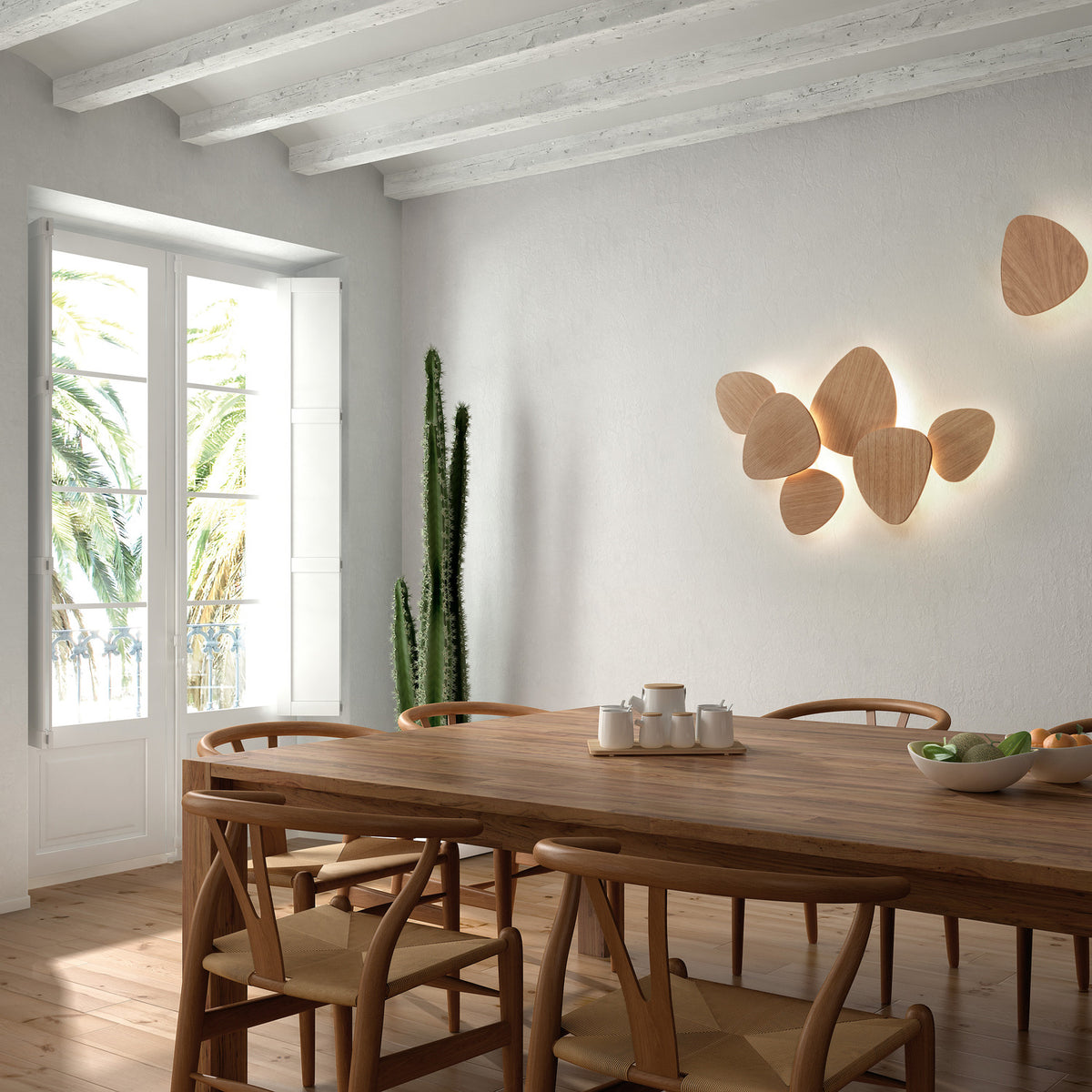 Tria 6 Light Wood LED Wall Light with pebble-shaped wood panels clustered on a white dining room wall, glowing softly behind each shape, with wishbone chairs and wooden table below.