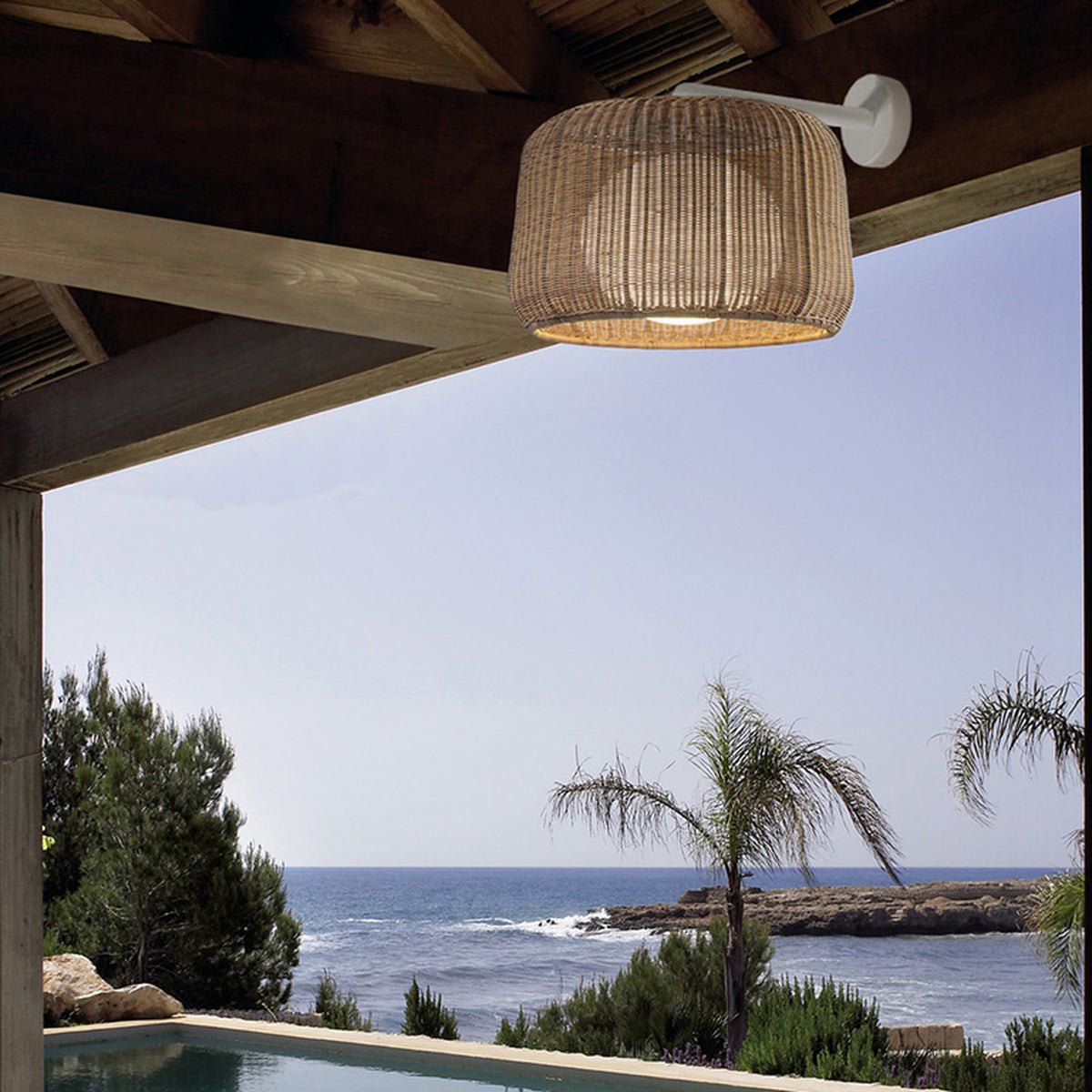 Fora A Drum LED Outdoor Wall Light in Light Beige woven drum shade on a slim white arm, shown mounted under a wooden pergola roof with a sea and palm tree view.