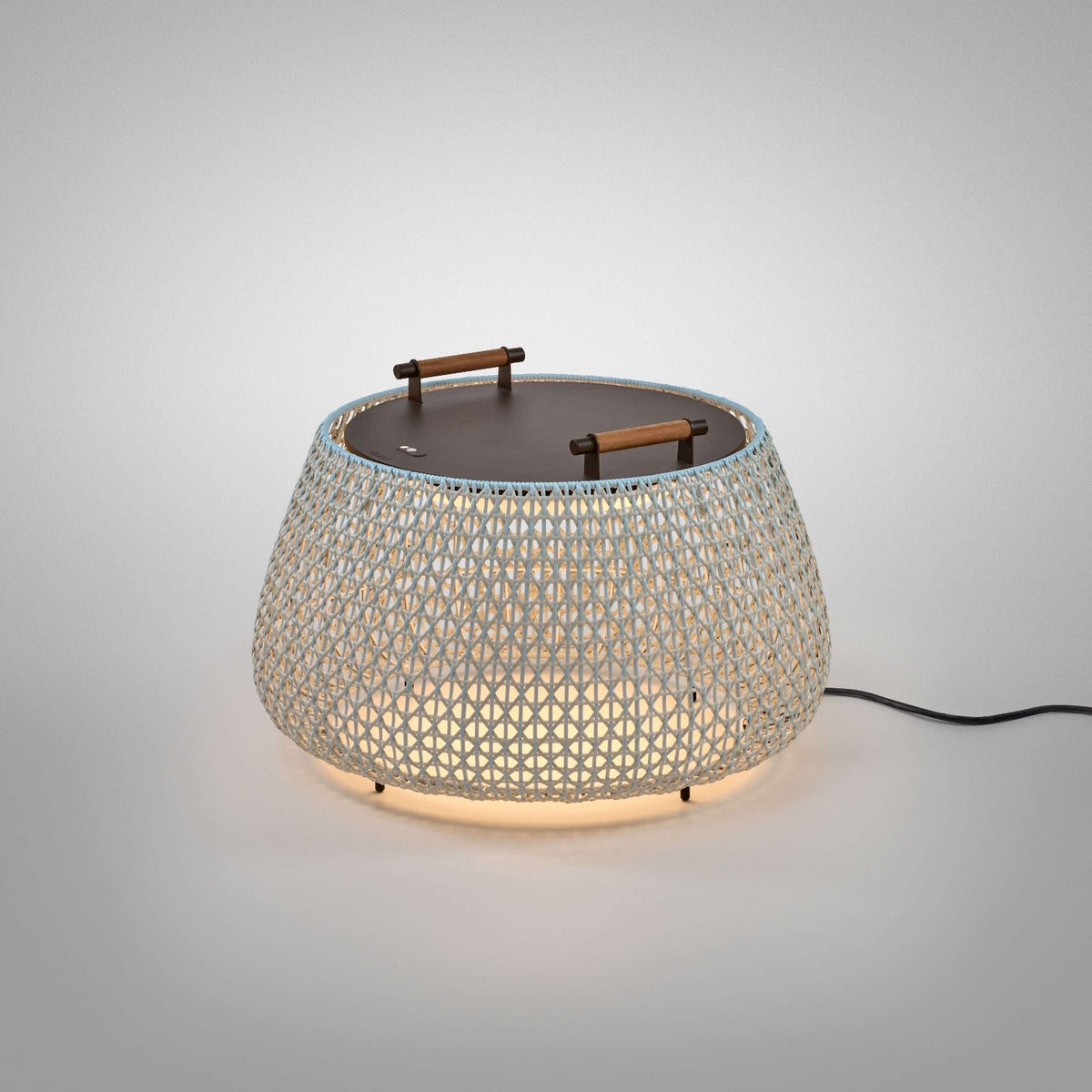 Nans Bag/38 outdoor floor lamp with a wide woven beige shade, flat dark top, and two teak carry handles, glowing softly against a plain light grey background.