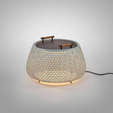 Nans Bag/38 outdoor floor lamp with a wide woven beige shade, flat dark top, and two teak carry handles, glowing softly against a plain light grey background.
