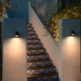 Nut A/01 black outdoor wall light with rounded bell-shaped head mounted on tiled staircase piers, casting warm LED downlight onto steps and greenery at night.