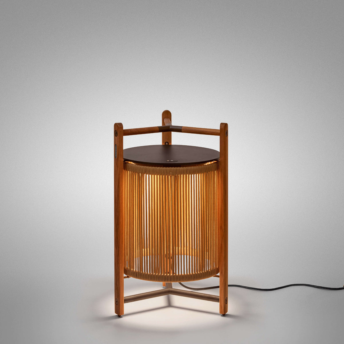 Kando P/76 outdoor floor lamp, lantern-style with sand nautical cord cylinder, three-legged teak wood frame, dark round lid and wooden handle, lit, on a plain light grey background.