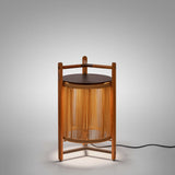 Kando P/76 outdoor floor lamp, lantern-style with sand nautical cord cylinder, three-legged teak wood frame, dark round lid and wooden handle, lit, on a plain light grey background.