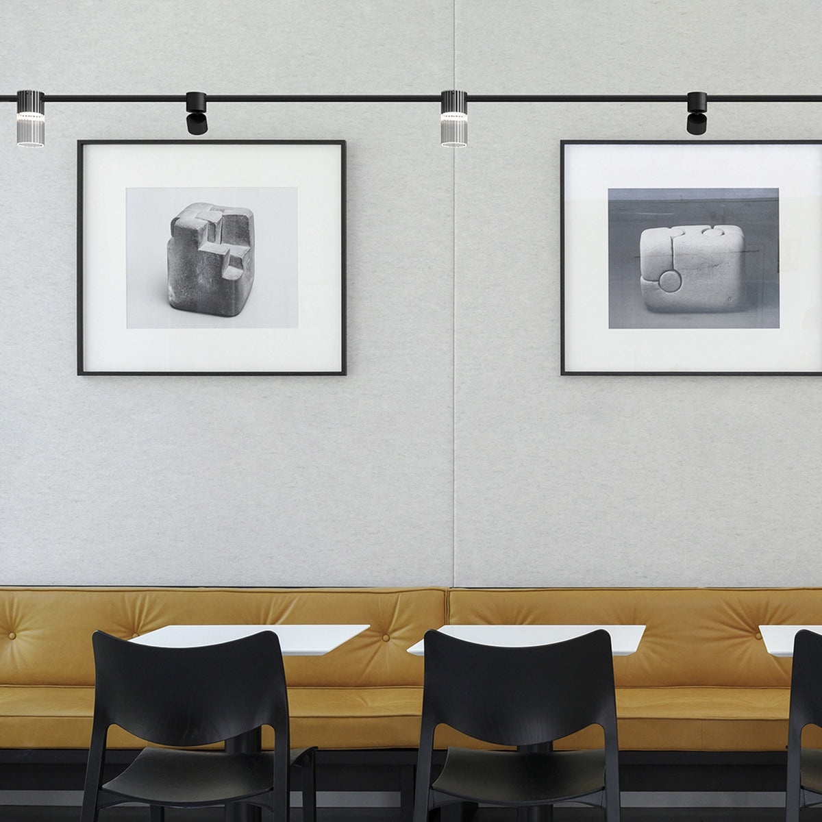 Black cylinder LED track spotlights mounted on ceiling rails, illuminating framed black-and-white artwork above a mustard leather banquette seating area.