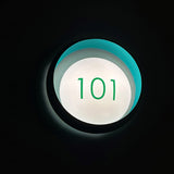 Ring W1 Number Glass Ring Wall Light in black with a teal-lit ring body and illuminated white front disc displaying the green number "101" against a dark background.