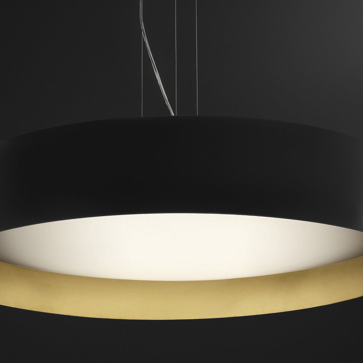 Ginevra 50 drum LED pendant light in matt black with a brass inner band, wide flat drum shape and glowing white diffuser underside, shown against a dark background.