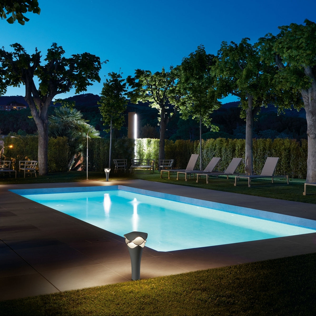 Cornet B/52 Cone LED bollard light in stone grey, shown glowing beside an illuminated outdoor pool at night with trees and loungers in the background.