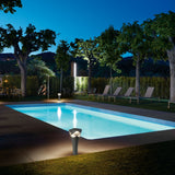 Cornet B/52 Cone LED bollard light in stone grey, shown glowing beside an illuminated outdoor pool at night with trees and loungers in the background.