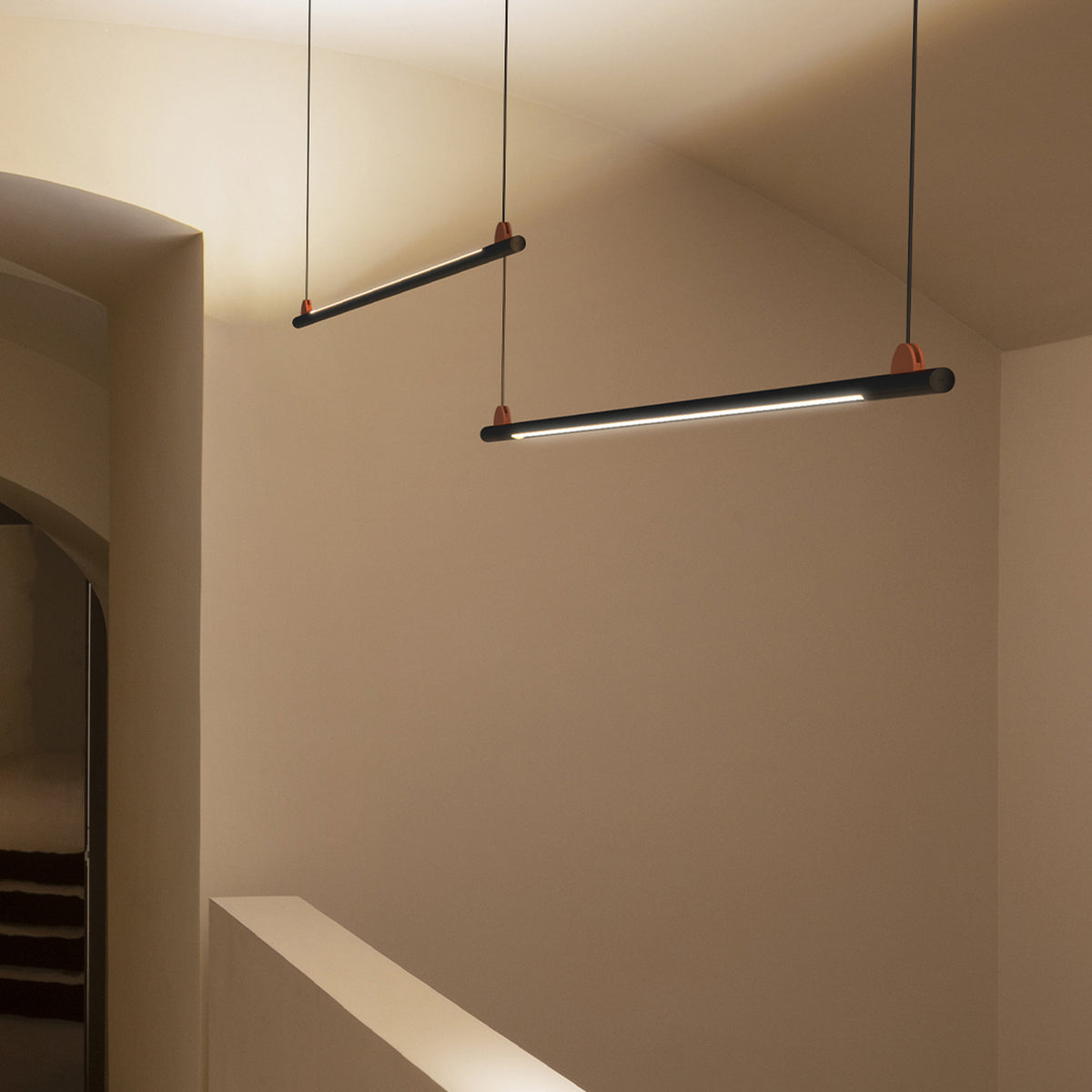 BIMU S150 Ele linear LED pendant lights in matte black with orange cable caps, hung from thin cables over a staircase landing, casting warm white light.