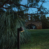 Nut B/60 Bell LED bollard light in textured black, slim square post with angled bell-shaped head, illuminated beside ornamental grasses in a garden at dusk.