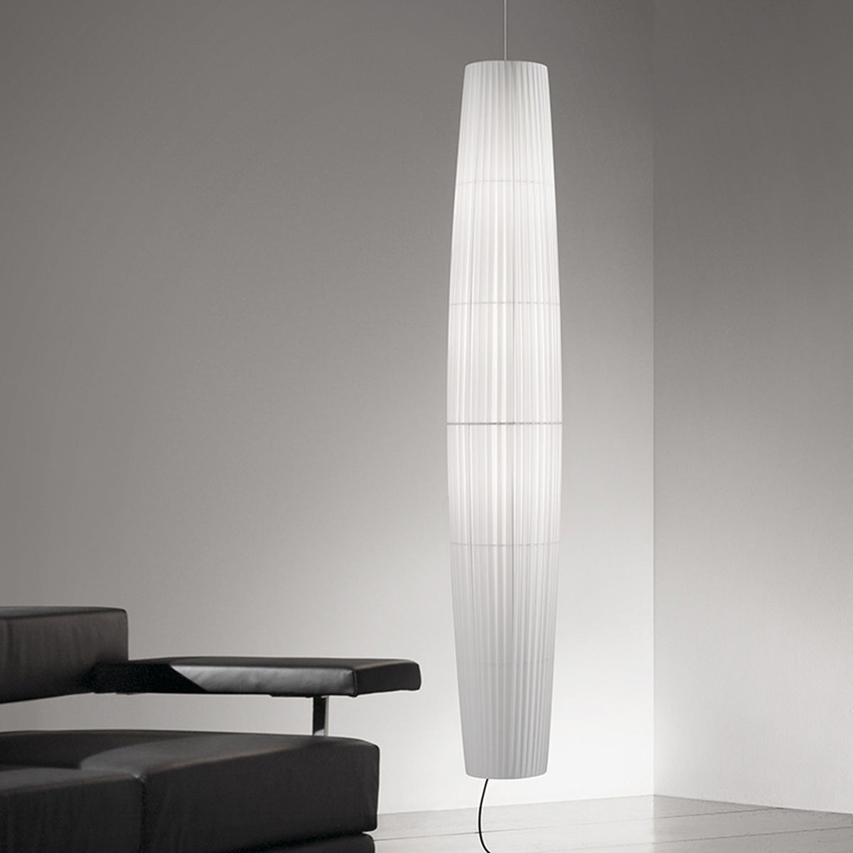 Maxi S/01 Ribbon LED pendant light with a tall, tapered white translucent fabric shade, pleated and banded, glowing in a modern living room beside a black leather sofa.