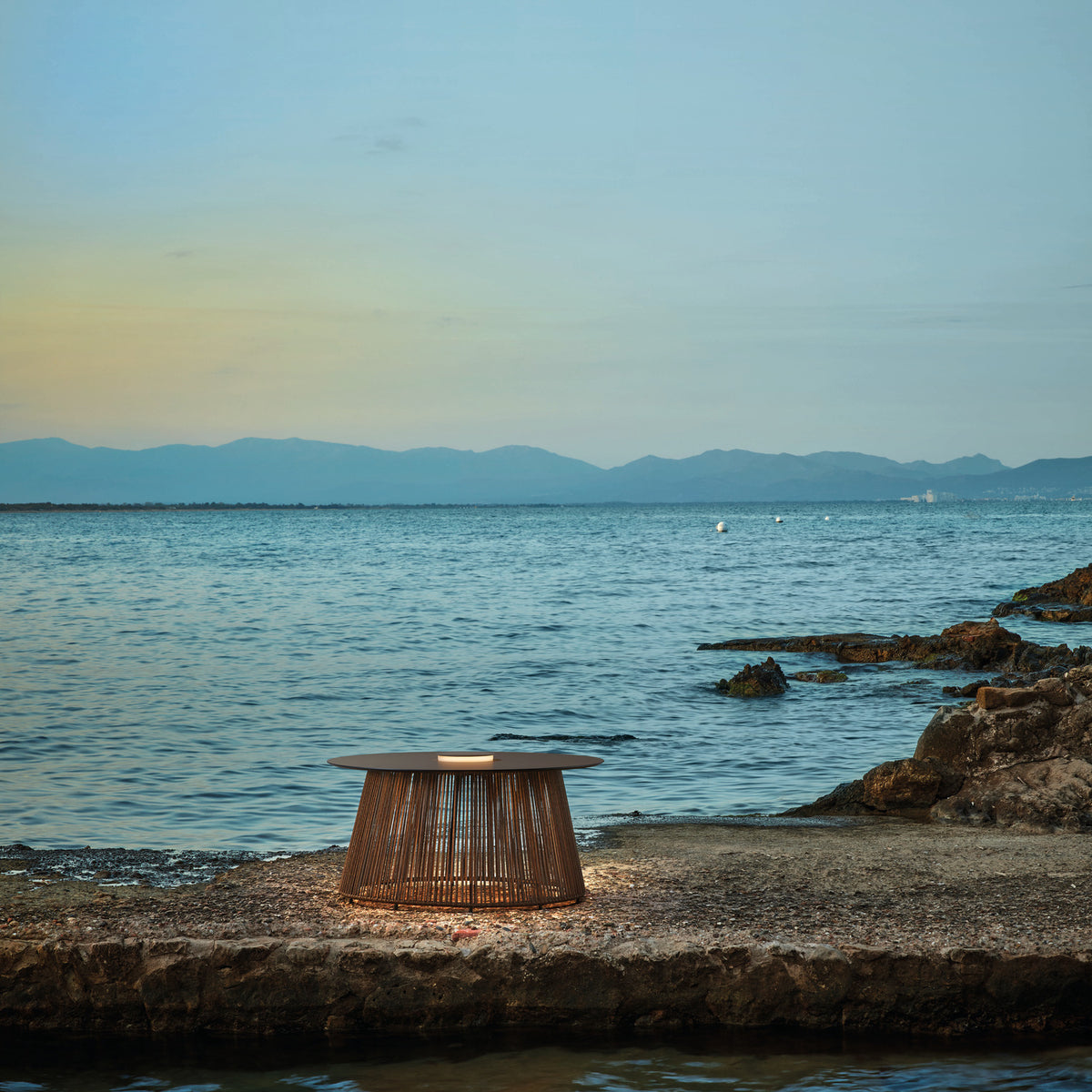 Nit/80/R rechargeable outdoor table lamp with round stone-finish top and woven brown synthetic fibre drum base, glowing centre light ring, placed on coastal rocks by the sea.