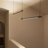 Two BIMU S100 Ele linear LED pendant lights in matte black with orange cable caps, hung level from thin cables in a neutral hallway with arched doorway.