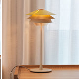 Overlay T25 table lamp with three stacked cone-shaped shades in beige over a glowing diffuser, slim stem, and round disc base, shown lit on a wooden table by a sheer curtain.