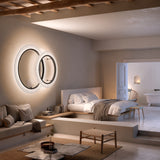 Roda PF/150 Ribbon LED wall light in white, two overlapping ring shades with black inner frames, glowing warm white above a bed in a softly lit bedroom.