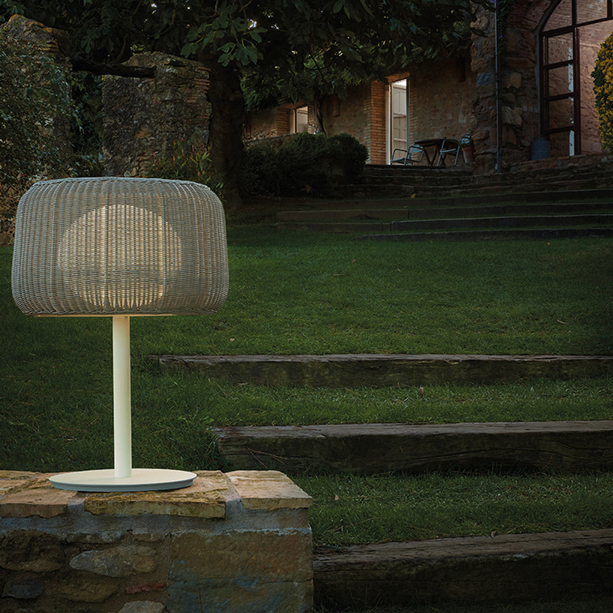 Fora M Drum LED Outdoor Table Lamp with a woven light beige drum shade, slim stem and round base, glowing on a stone wall beside a garden lawn at dusk.
