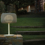 Fora M Drum LED Outdoor Table Lamp with a woven light beige drum shade, slim stem and round base, glowing on a stone wall beside a garden lawn at dusk.