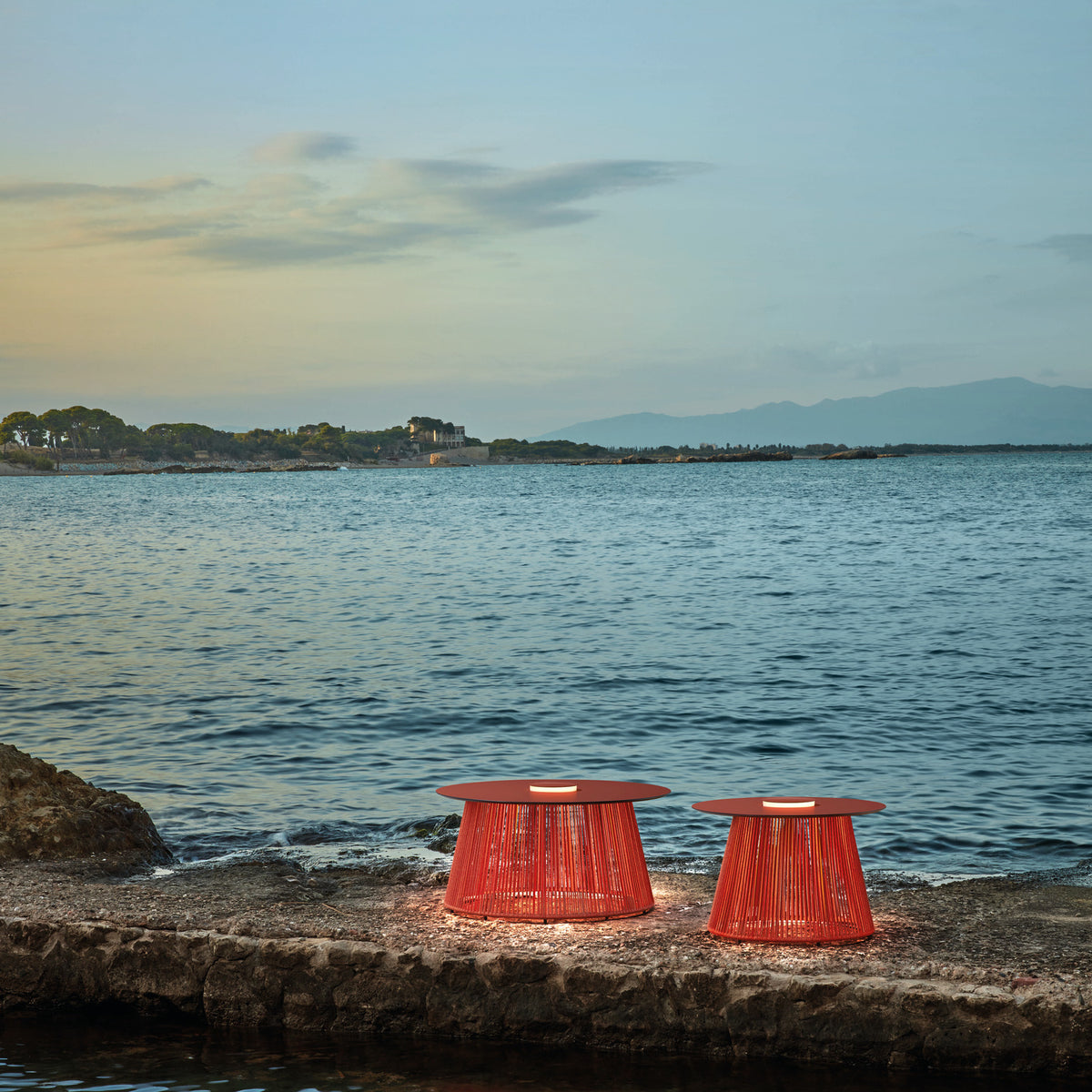 Two Nit/60/R woven rechargeable outdoor table lamps in orange, flared cord bodies with round tops and integrated LED light rings, set on a coastal stone ledge by the sea.