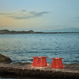 Two Nit/60/R woven rechargeable outdoor table lamps in orange, flared cord bodies with round tops and integrated LED light rings, set on a coastal stone ledge by the sea.