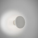 Arid A/01 round LED wall light in aluminium with Textured White finish, fluted disc face on a short stem, glowing softly against a plain white wall.
