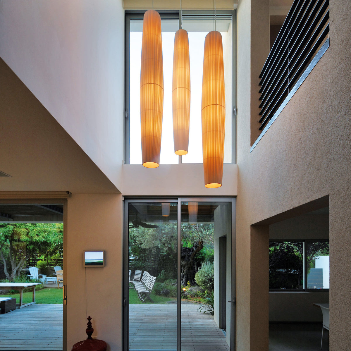 Three Maxi S/01 Ribbon LED pendant lights with tall, slender cream pleated shades hanging in a double-height hallway above a staircase and glass doors.