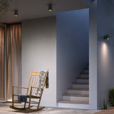 Nut PF/10 small bell-shaped outdoor ceiling light in black, mounted on a grey wall above a staircase in an outdoor seating area with a wooden rocking chair.