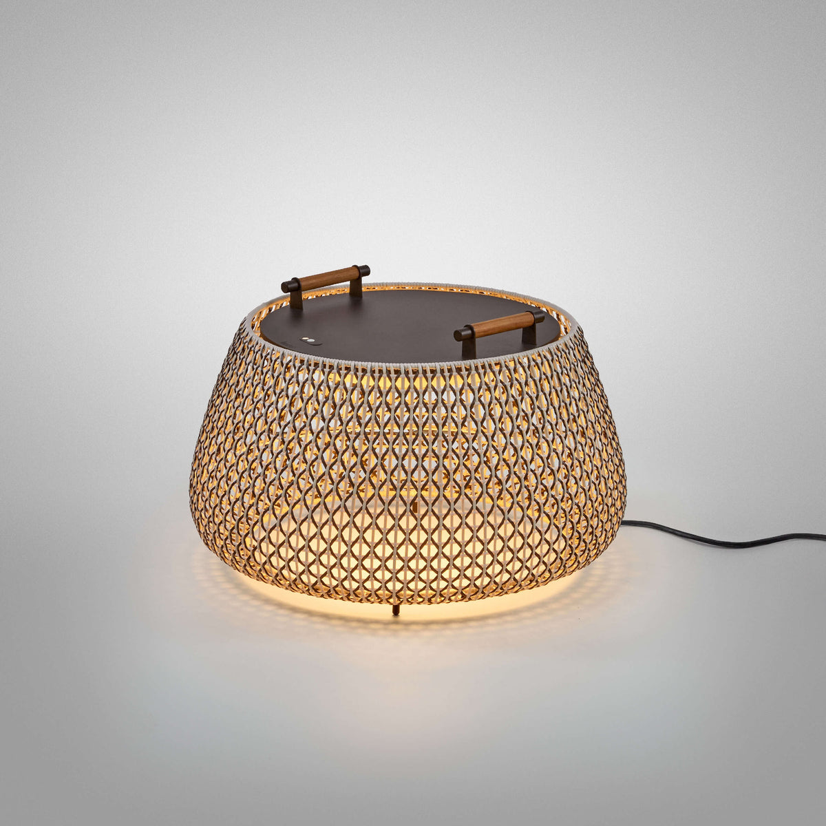 Nans Bag/38 Wood LED outdoor floor lamp with woven brown lattice shade, dark flat top, two teak handles, illuminated against a plain grey background.