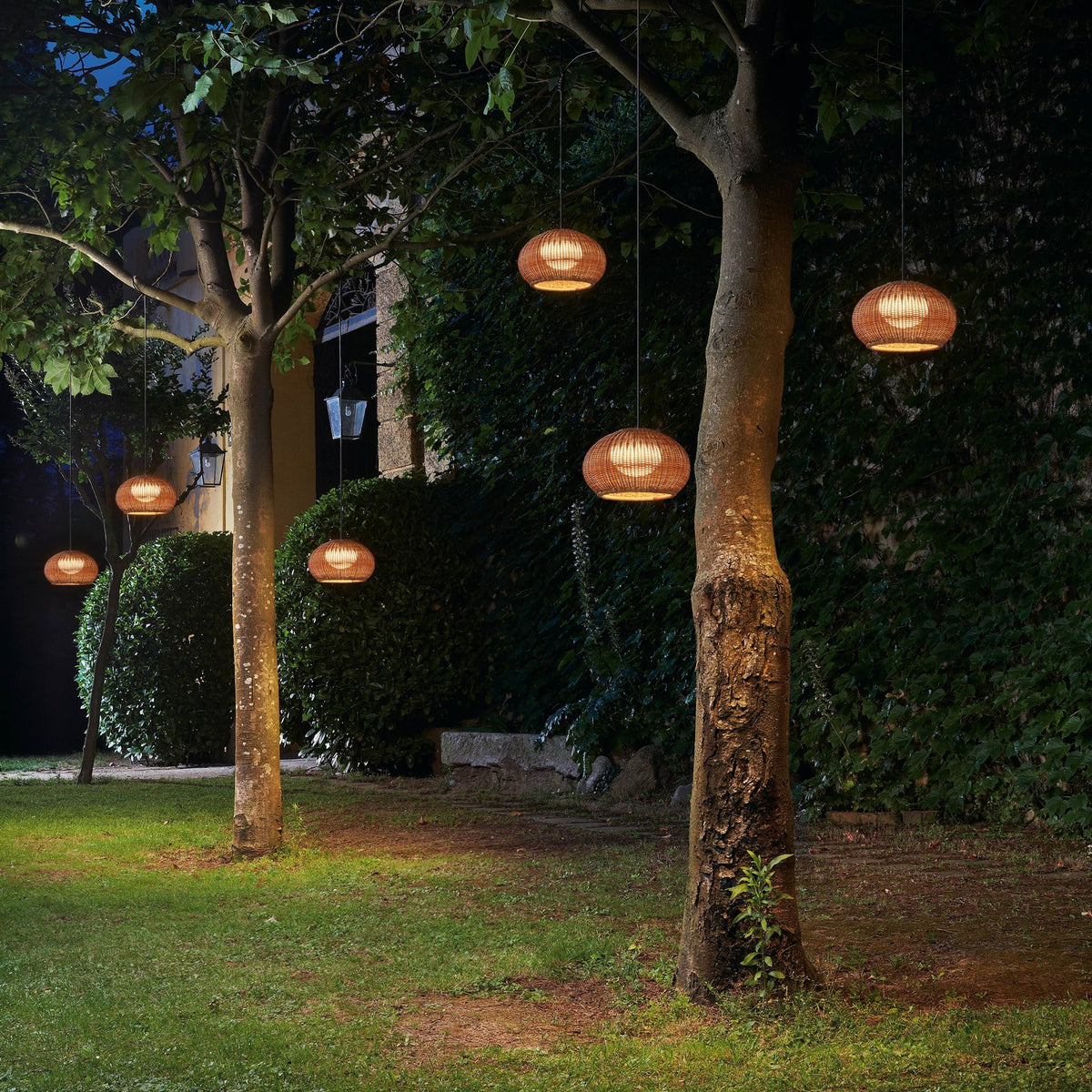 Garota Hang Globe LED outdoor pendant lights with brown woven globe shades hung from trees, glowing warmly in a garden setting at night.