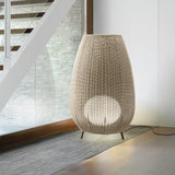 Amphora 03 Woven LED Outdoor Floor Lamp in beige, a tall vase-shaped woven shade on tapered legs, glowing softly, shown beside a staircase and glass panel indoors.