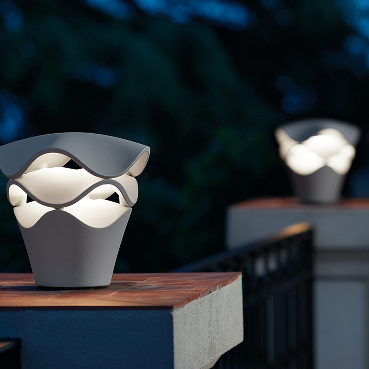 Cornet B/27 Cone LED bollard light in Stone Grey, tapered cone base with stacked wavy layers glowing white, shown outdoors on a wooden ledge at dusk.